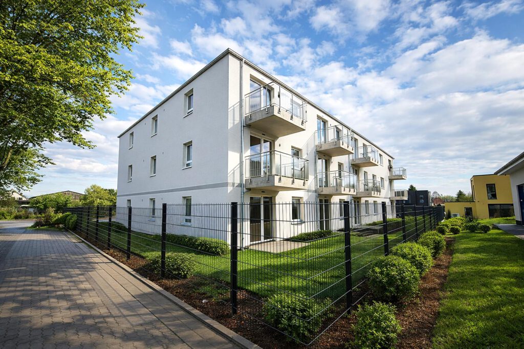 Außenansicht des Mehrfamilienhauses am Werbeker Ring in Pattensen mit weißer Putzfassade, Balkonen und gepflegten Grünanlagen – geförderter Neubau mit 24 Wohneinheiten, KfW-55-Standard, Fertigstellung 2026