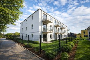 Außenansicht des Mehrfamilienhauses am Werbeker Ring in Pattensen mit weißer Putzfassade, Balkonen und gepflegten Grünanlagen – geförderter Neubau mit 24 Wohneinheiten, KfW-55-Standard, Fertigstellung 2026